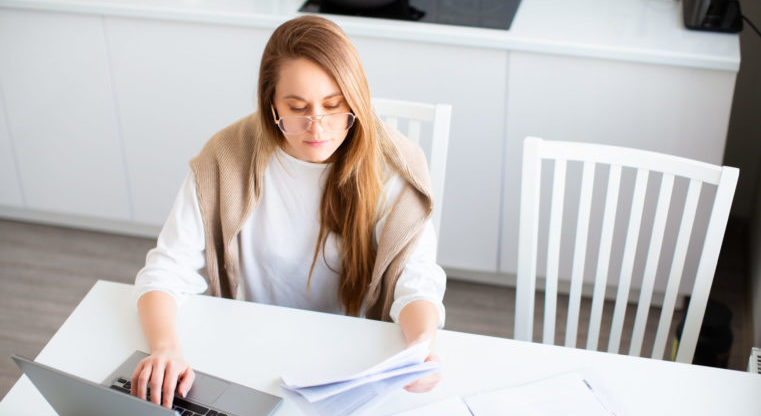 Online work concept. Woman works online in front of a laptop monitor. She fills out tax forms or pays bills. Photo in home interior. Top view.